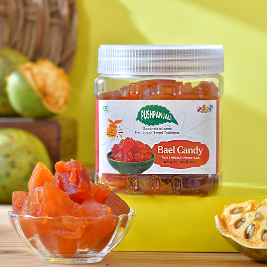 A jar of Pushpanjali Bael Candy is displayed on a yellow surface with a glass bowl of orange-red bael pieces in the foreground and bael fruits in the background, creating a vibrant, natural product presentation.