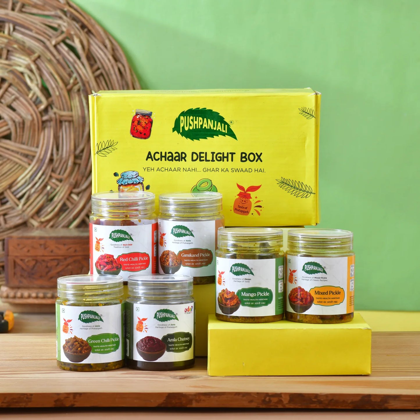 A Pushpanjali Achaar Delight Box set displayed on a wooden surface against a green wall. The set includes a large yellow box and six pet jars of various pickles and chutney, arranged neatly with a yellow block as a stand.