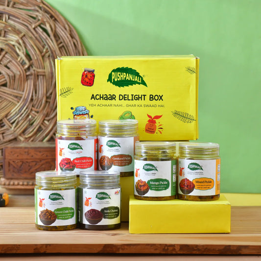 A Pushpanjali Achaar Delight Box set displayed on a wooden surface against a green wall. The set includes a large yellow box and six pet jars of various pickles and chutney, arranged neatly with a yellow block as a stand.
