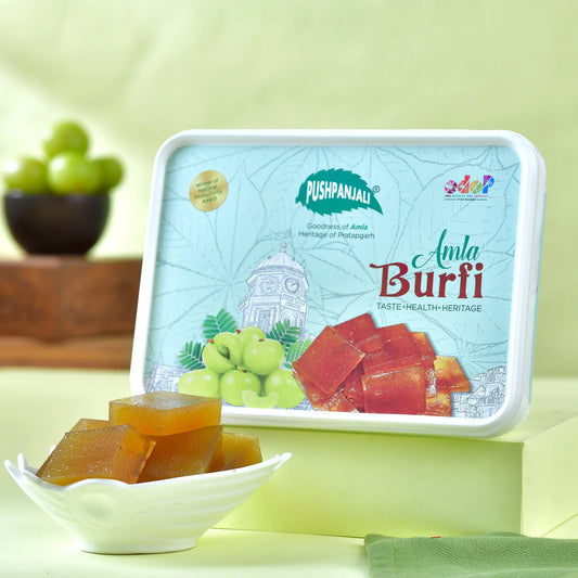A plastic box of Pushpanjali Amla Burfi sweets is displayed on a light green pedestal with several pieces of the amber-brown sweets in a small white dish in the foreground. The plastic box features a light blue and white design with illustrations of a amla fruit, and the product name.