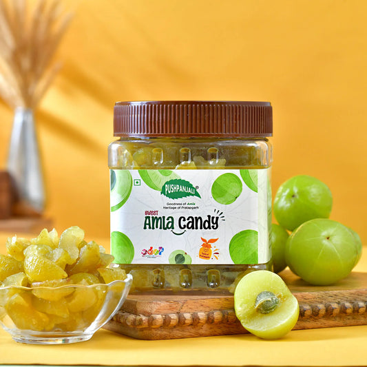 A jar of Pushpanadi Amla Candy is displayed on a wooden board with fresh green amlas, a small glass bowl of yellow candied amlas, and a yellow background.