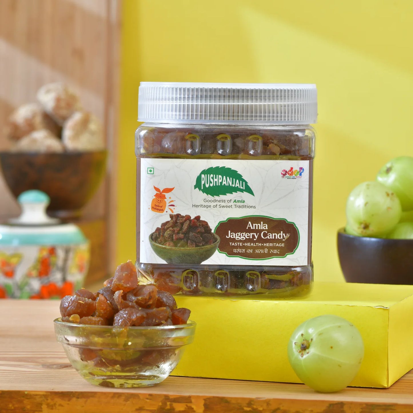 A jar of Pushpanjali Amla Jaggery Candy placed on a wooden surface with loose amlas and a yellow box around it, against a bright yellow background.