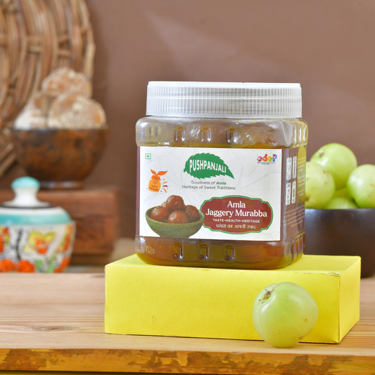 A jar of Pushpanjali Amla Jaggery Murabba (sweet jaggery) displayed on a yellow block on a wooden table, with whole green grapes in the background.