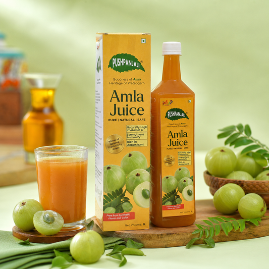 A bottle and box of Pushpanjali Amla Juice are displayed on a wooden board with whole amlas, a glass of amla juice, and green leaves against a soft green background.
