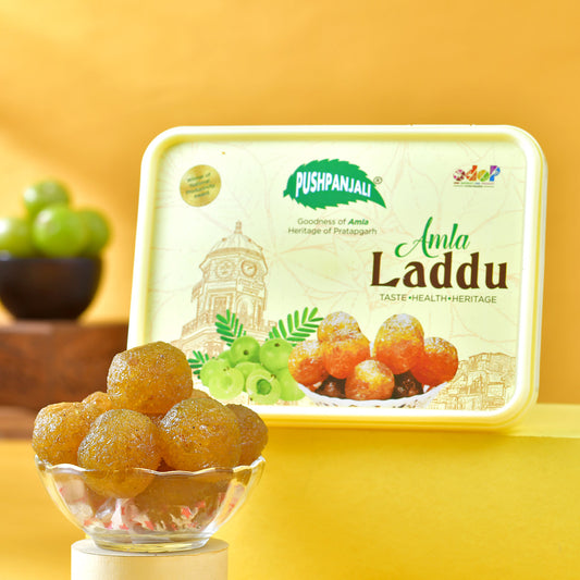 A plastic box of Pushpanjali Amla Laddu sweets displayed next to a glass bowl filled with golden-orange amla laddu balls, set against a bright yellow background.
