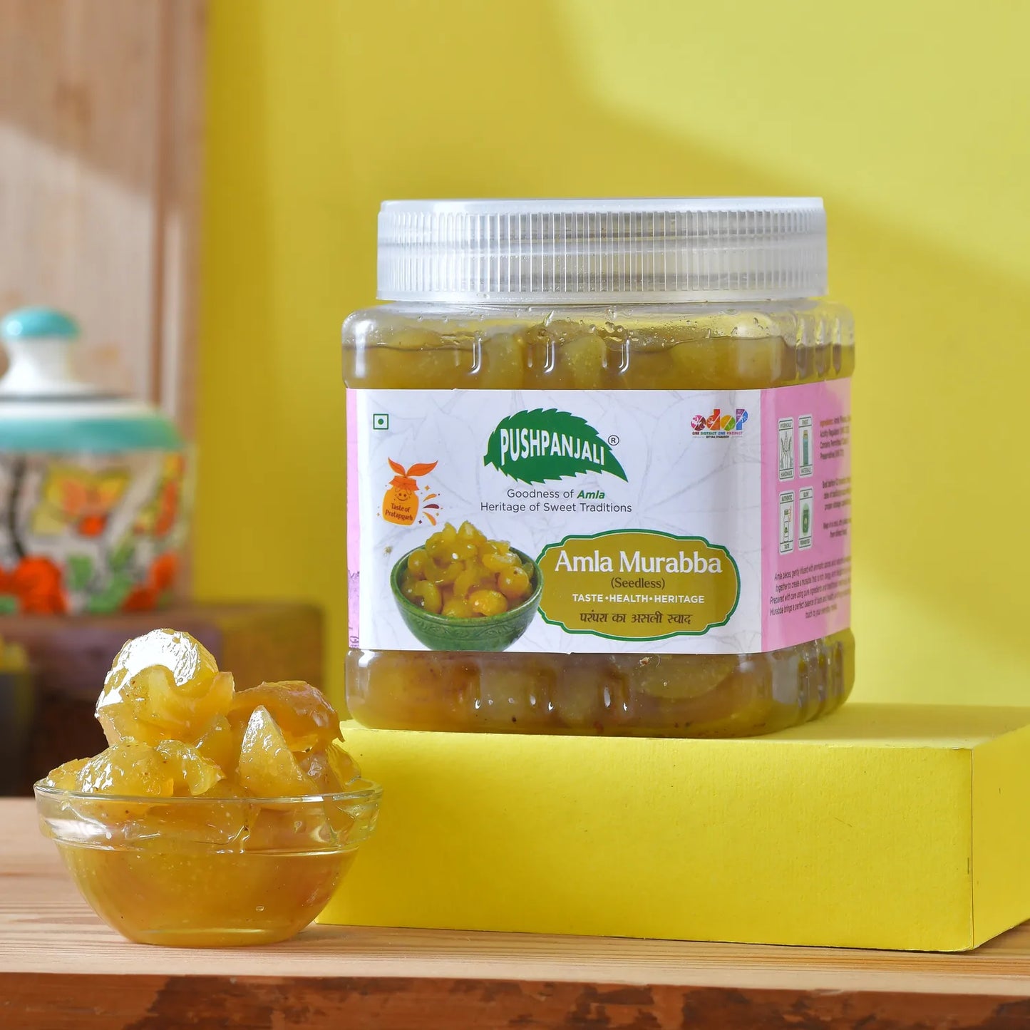A jar of Pushpanjali Amla Murabba (pitted amla murabba) displayed on a yellow block with a small glass bowl of glistening amla pieces in the foreground, set against a bright yellow background.