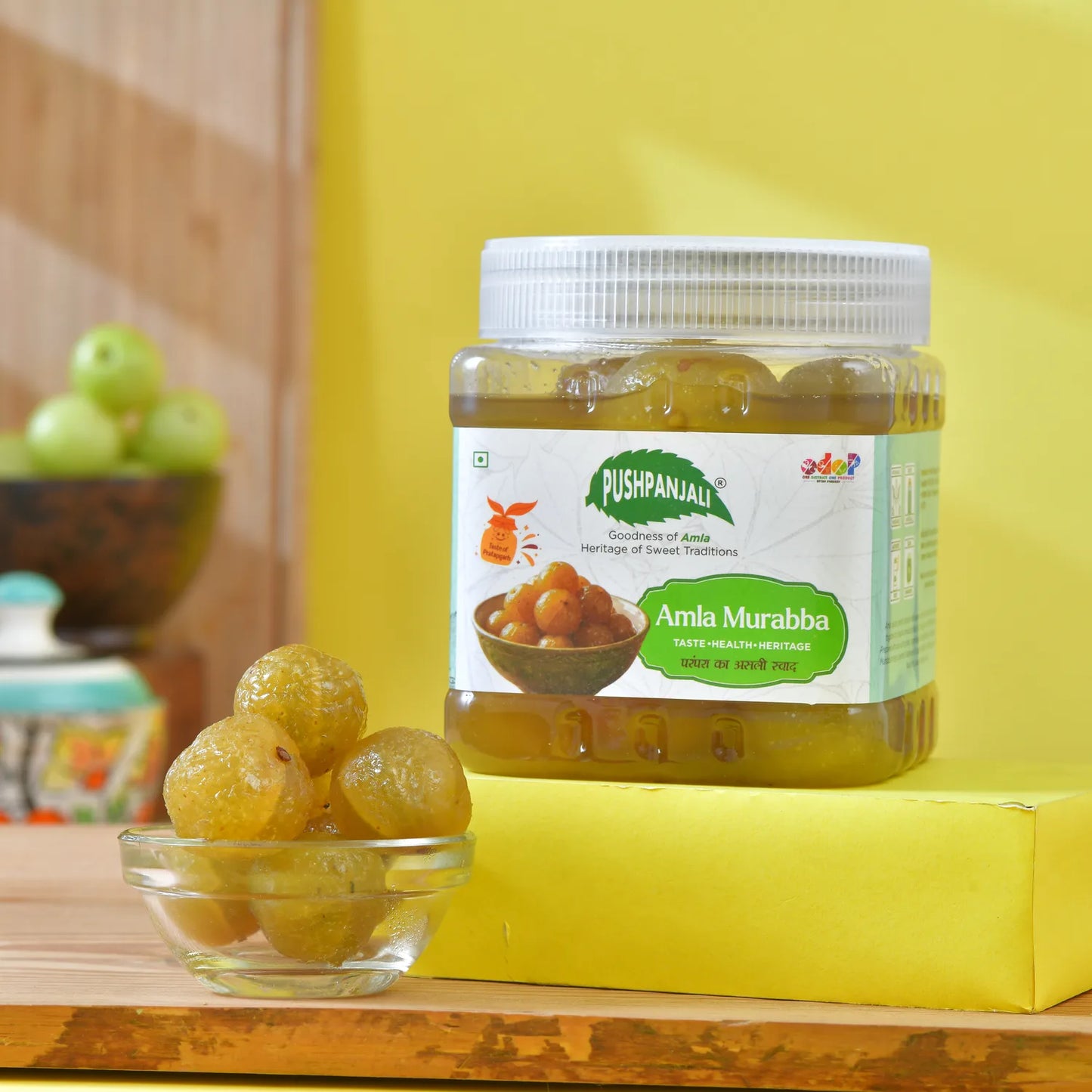 A jar of Pushpanjali Amla Murabba sits on a yellow block next to a small glass bowl of whole amlas, with a blurred background showing more amlas.