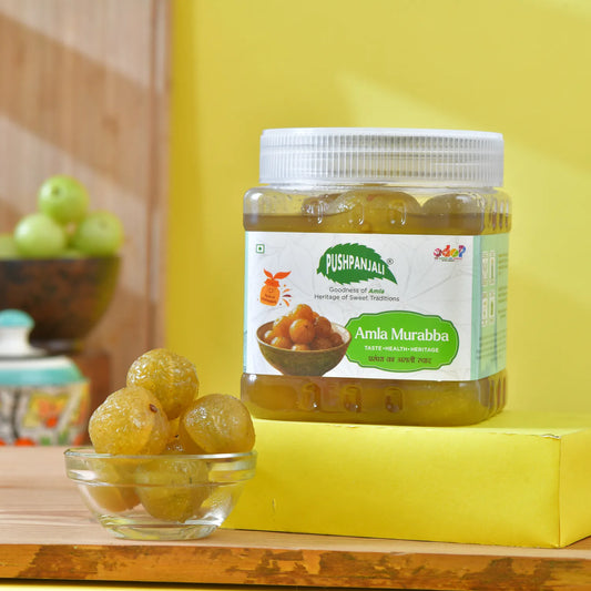A jar of Pushpanjali Amla Murabba sits on a yellow block next to a small glass bowl of whole amlas, with a blurred background showing more amlas.