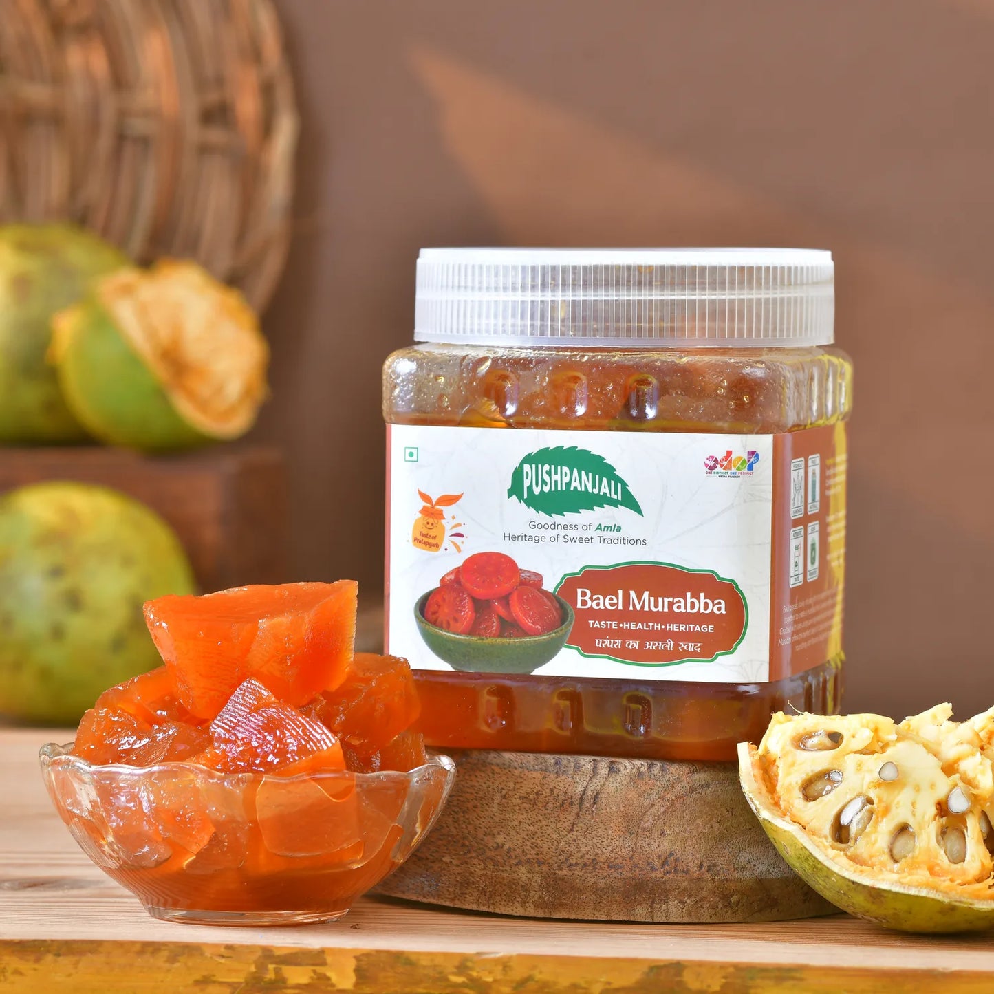 A jar of Pushpanjali Bael Murabba on a wooden stand, with a glass bowl of chunky orange-red bael murabba and fresh bael halves in the foreground against a warm brown background.