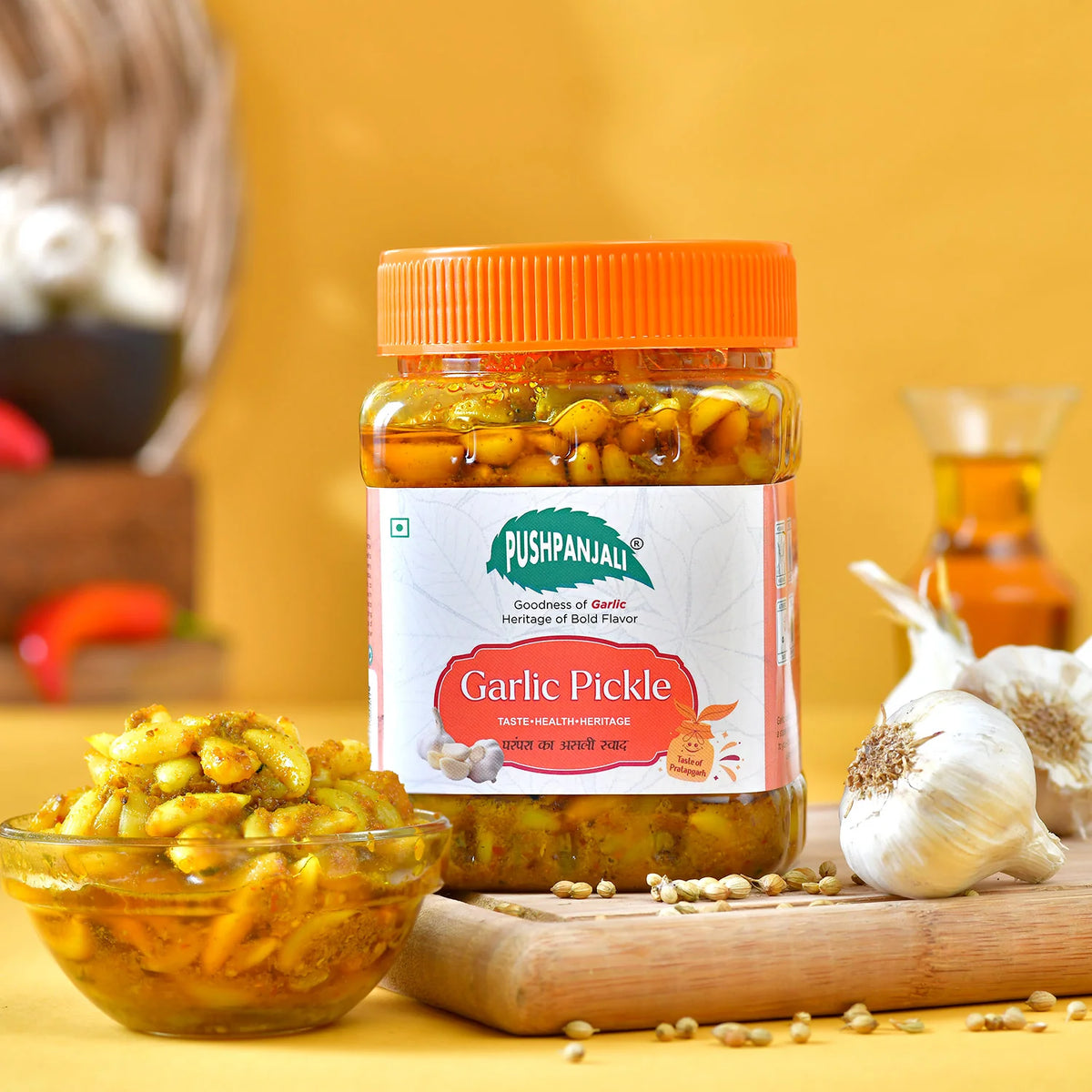 A jar of Pushpanjali Garlic Pickle with an orange lid and white label, displayed on a wooden board with garlic bulbs, a small bowl of pickled garlic, and a blurred background with warm yellow tones.