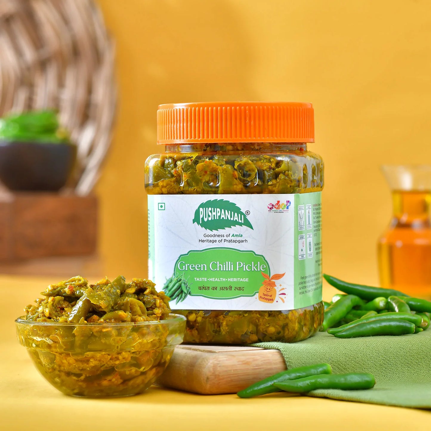 A jar of Pushpanjali Green Chilli Pickle with an orange lid, displayed on a yellow background with a glass bowl of pickle, green chilies, and a wooden spoon.
