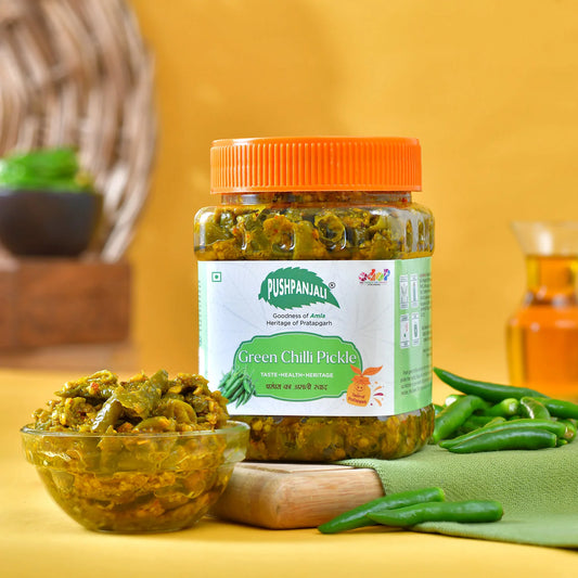 A jar of Pushpanjali Green Chilli Pickle with an orange lid, displayed on a yellow background with a glass bowl of pickle, green chilies, and a wooden spoon.