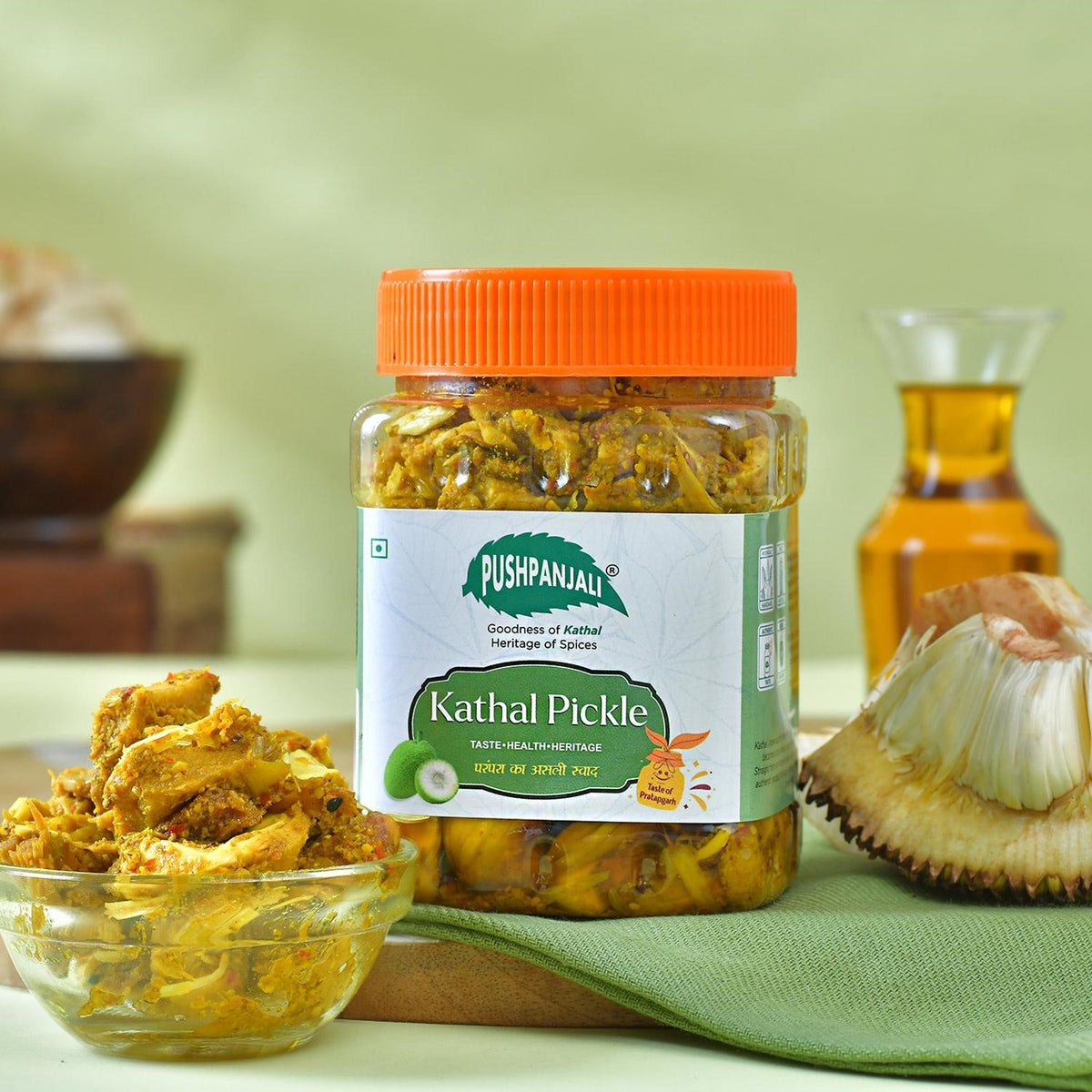 A jar of Pushpanjali Kathal Pickle with a bright orange lid, set on a green cloth with a bowl of pickle, jackfruit, and a bottle of oil in a styled background.