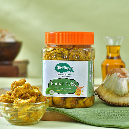 A jar of Pushpanjali Kathal Pickle with a bright orange lid, set on a green cloth with a bowl of pickle, jackfruit, and a bottle of oil in a styled background.
