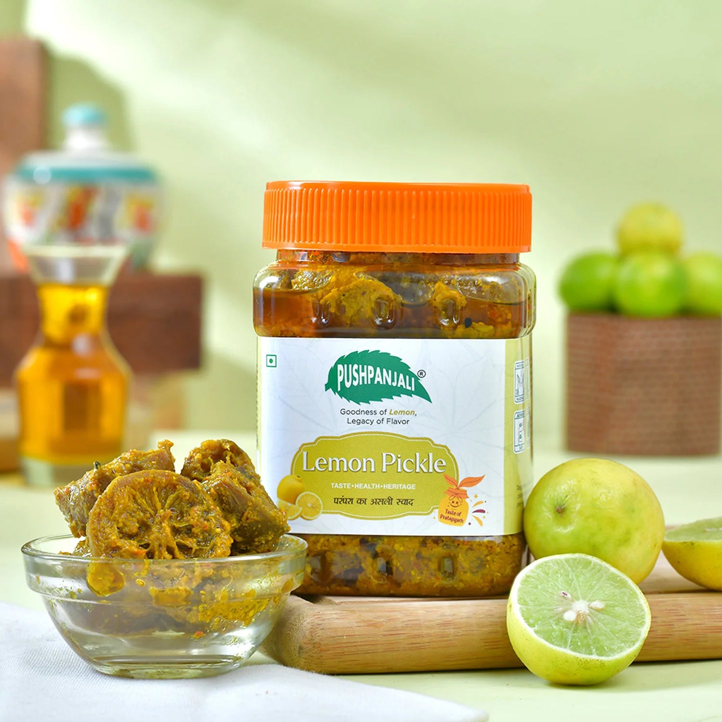 A jar of Pushpanjali Lemon Pickle with an orange lid and white label, styled on a wooden board with whole and halved limes and a small glass bowl of pickle slices in the foreground, with a softly blurred kitchen background.
