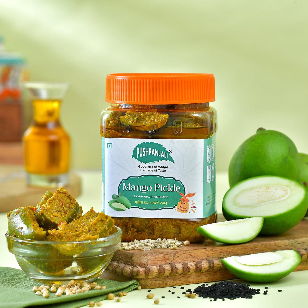A jar of Pushpanjali Mango Pickle is displayed on a wooden cutting board with fresh mango slices, a small glass bowl of pickle, and spices, set against a soft green background.