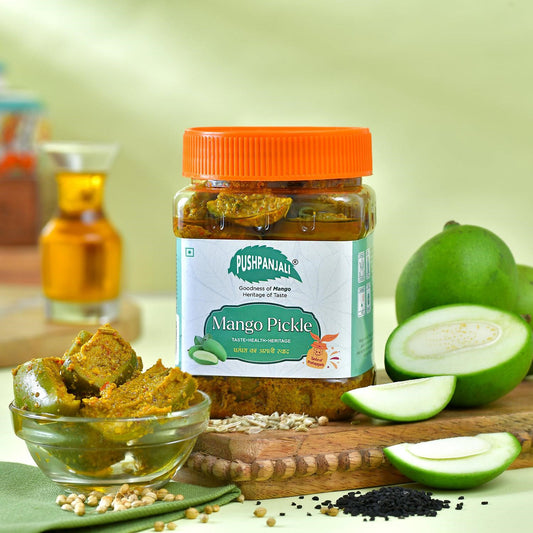 A jar of Pushpanjali Mango Pickle is displayed on a wooden cutting board with fresh mango slices, a small glass bowl of pickle, and spices, set against a soft green background.