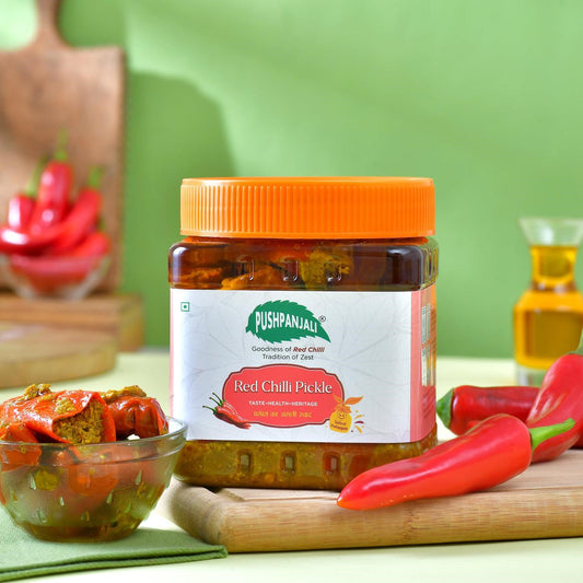 A jar of Pushpanjali Red Chilli Pickle with an orange lid, set on a wooden cutting board with fresh red chilies, a small bowl of pickled vegetables, and a green background.